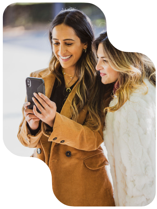 Two people smiling while looking at a smartphone. One is wearing a brown jacket and the other a white coat.