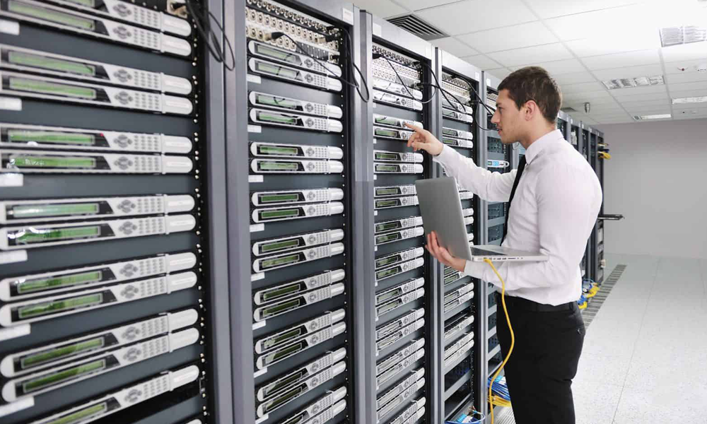 A person in a white shirt stands in a server room, holding a laptop and adjusting equipment on a rack.