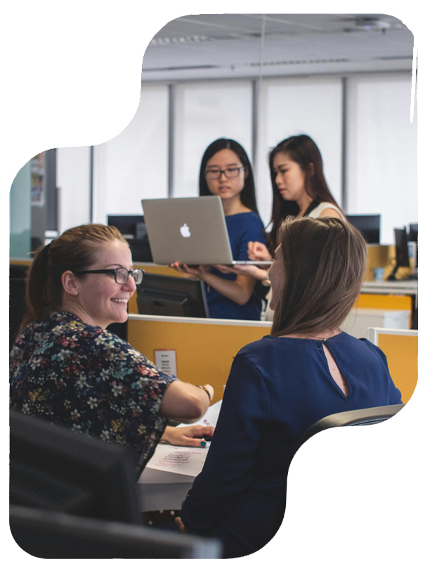 Four women collaborate in an office, two seated at a desk and two standing with a laptop.