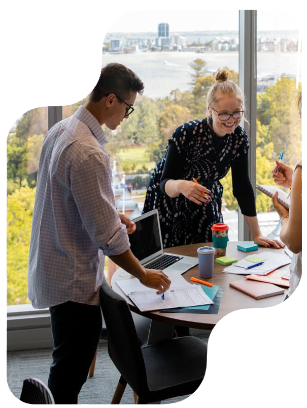 Three people working together at a round table in a bright office with large windows and a view of the city and trees. One is standing, another is seated, and they are surrounded by paperwork.