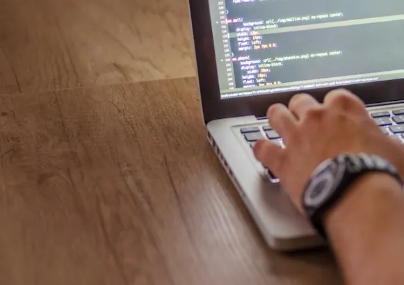 A person types on a laptop keyboard displaying a code editor, highlighting why professional IT consulting is essential for business growth, all on a wooden surface.