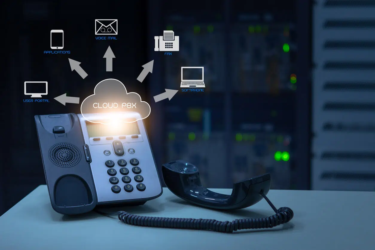 Office phone with a detached handset on a table, displaying a cloud PBX graphic linking to icons for voicemail, fax, applications, software, and user profile. Servers are visible in the background.