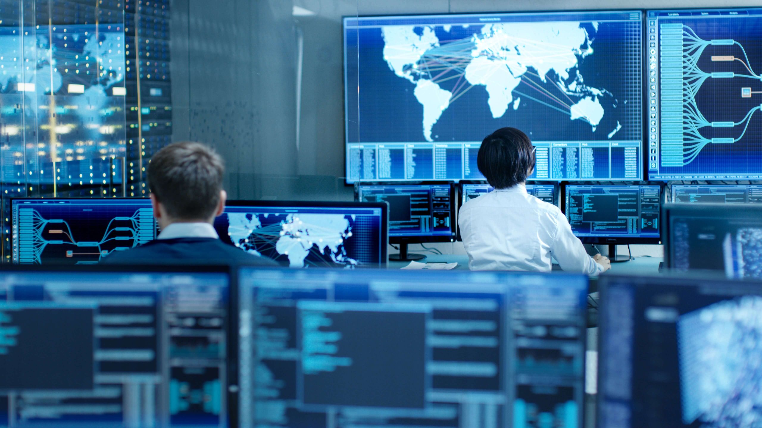 People in a control room monitor global data on multiple screens displaying world maps and information.