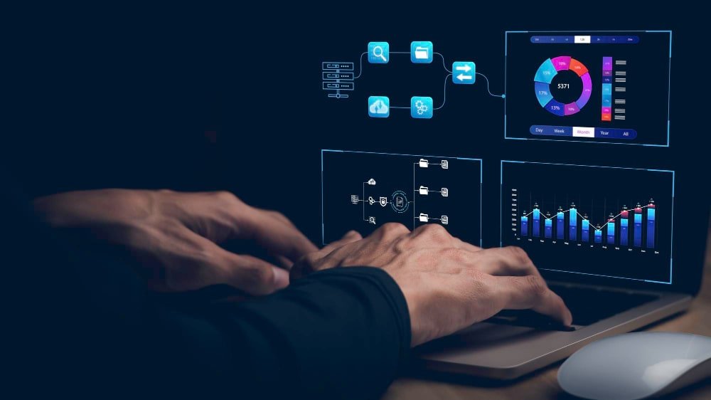 Hands typing on a laptop with various data and graphs displayed on screens in a dark setting.