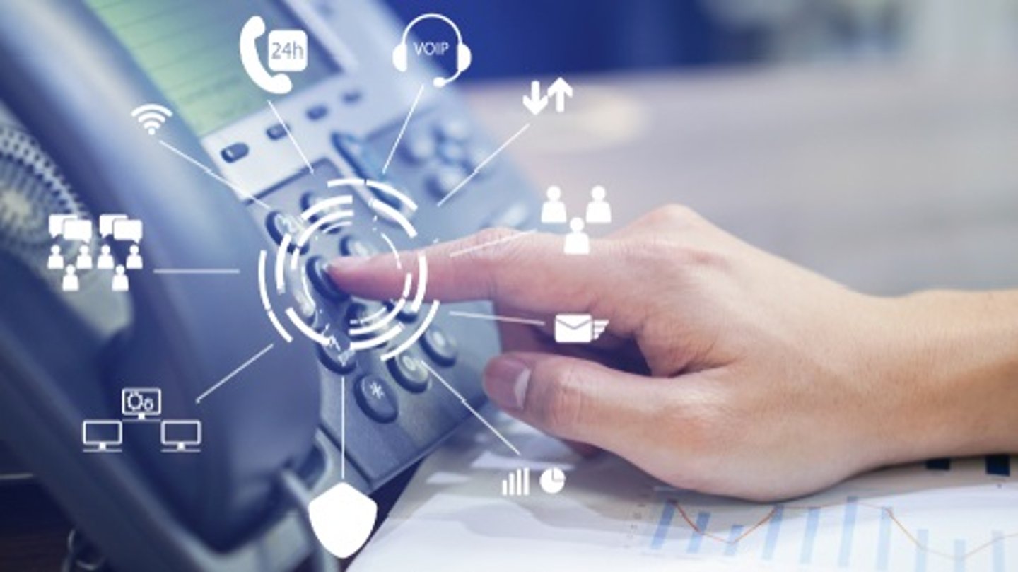 A hand pressing a button on a VOIP office phone, surrounded by digital communication icons, on a desk with a paper chart.