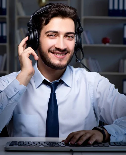 A man in a white shirt and dark tie, wearing a headset, sits at a desk with a keyboard, smiling at the camera. Bookshelves are visible in the background.