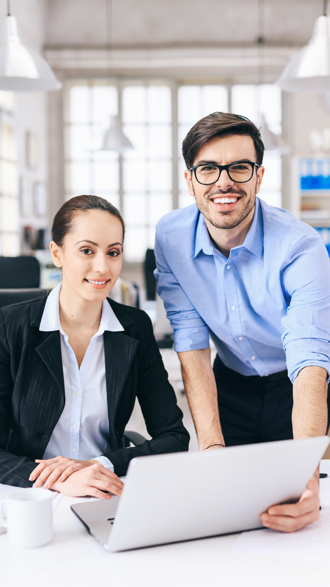 Two colleagues in professional attire smiling at a desk with a laptop in an office setting.