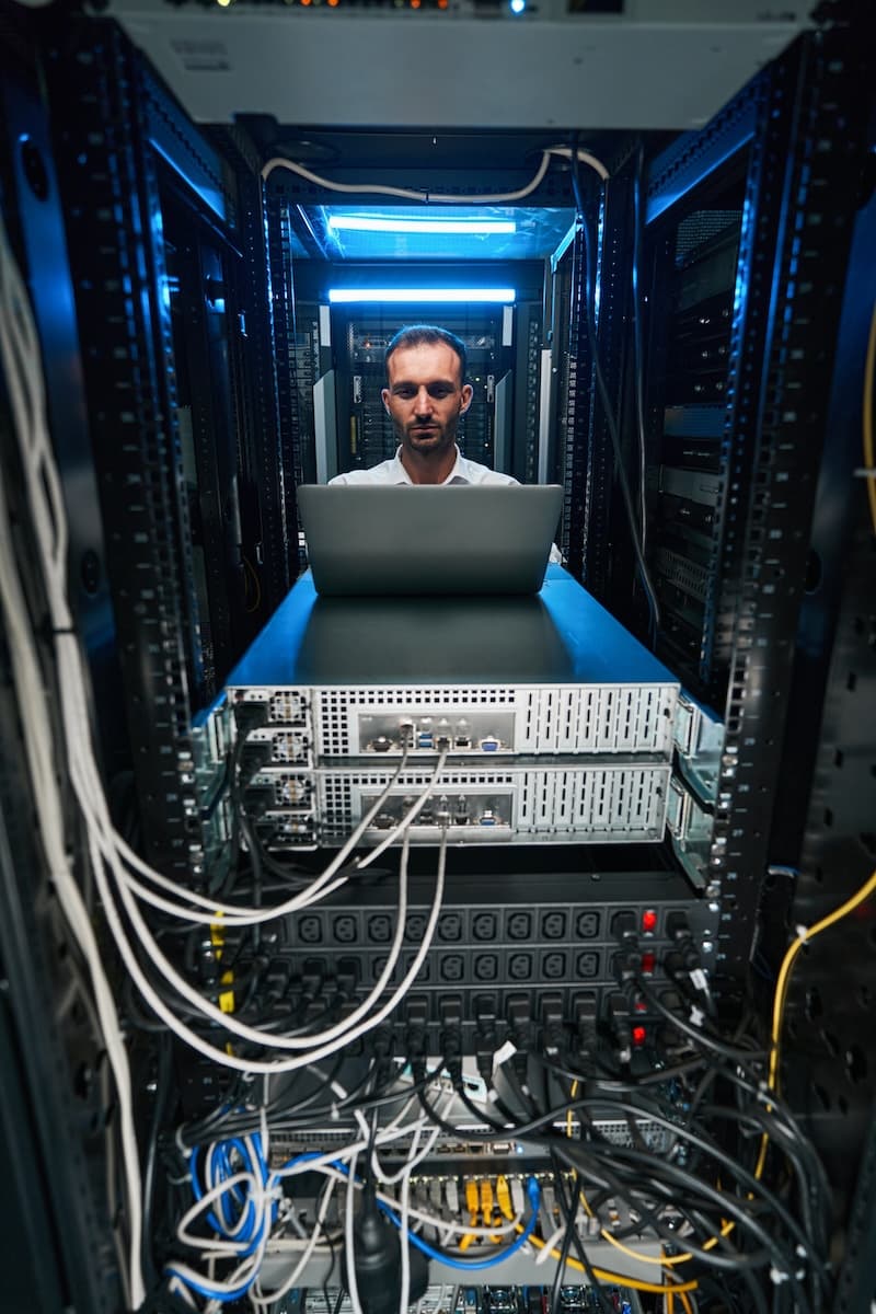 A person stands in a server room, meticulously working on a laptop surrounded by racks of servers and network cables, ensuring the new data is properly integrated into the system.