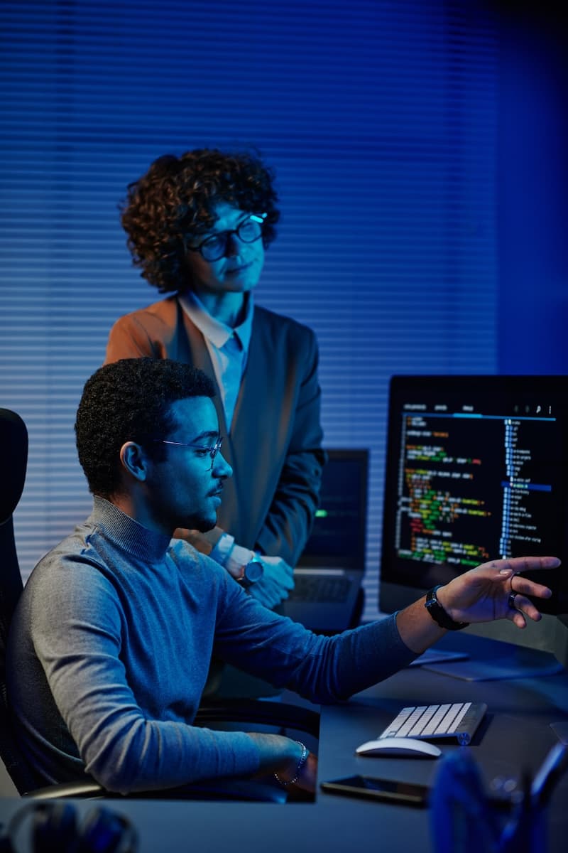 In a dimly lit office with blue tones, two people meticulously review code on a computer screen, analyzing new data updates.