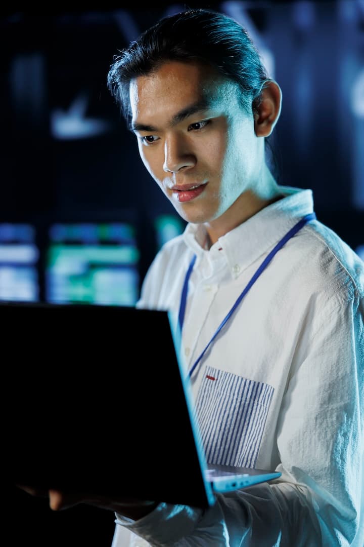 Person in a white shirt looking at a laptop screen, with blurred background featuring multiple monitors.