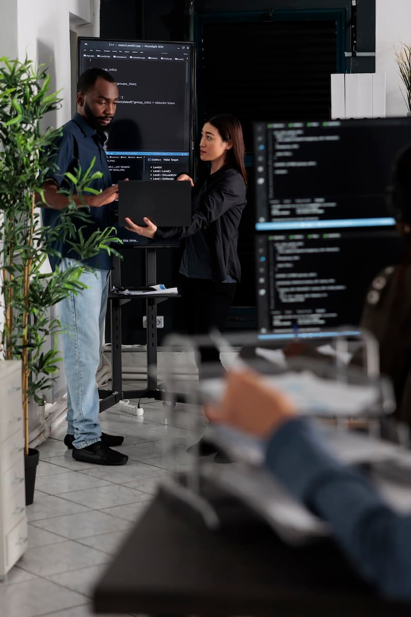 Two people stand near a computer monitor displaying code, holding a laptop. Another person is seated in the foreground, working at a desk with two monitors.