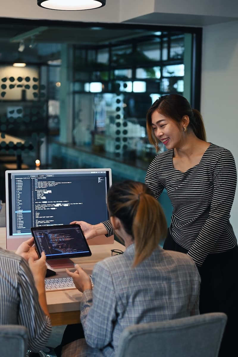 Three people collaborate in an office. One person stands, smiling, while two others sit, analyzing new data on a tablet and a computer screen displaying code for the backup page.