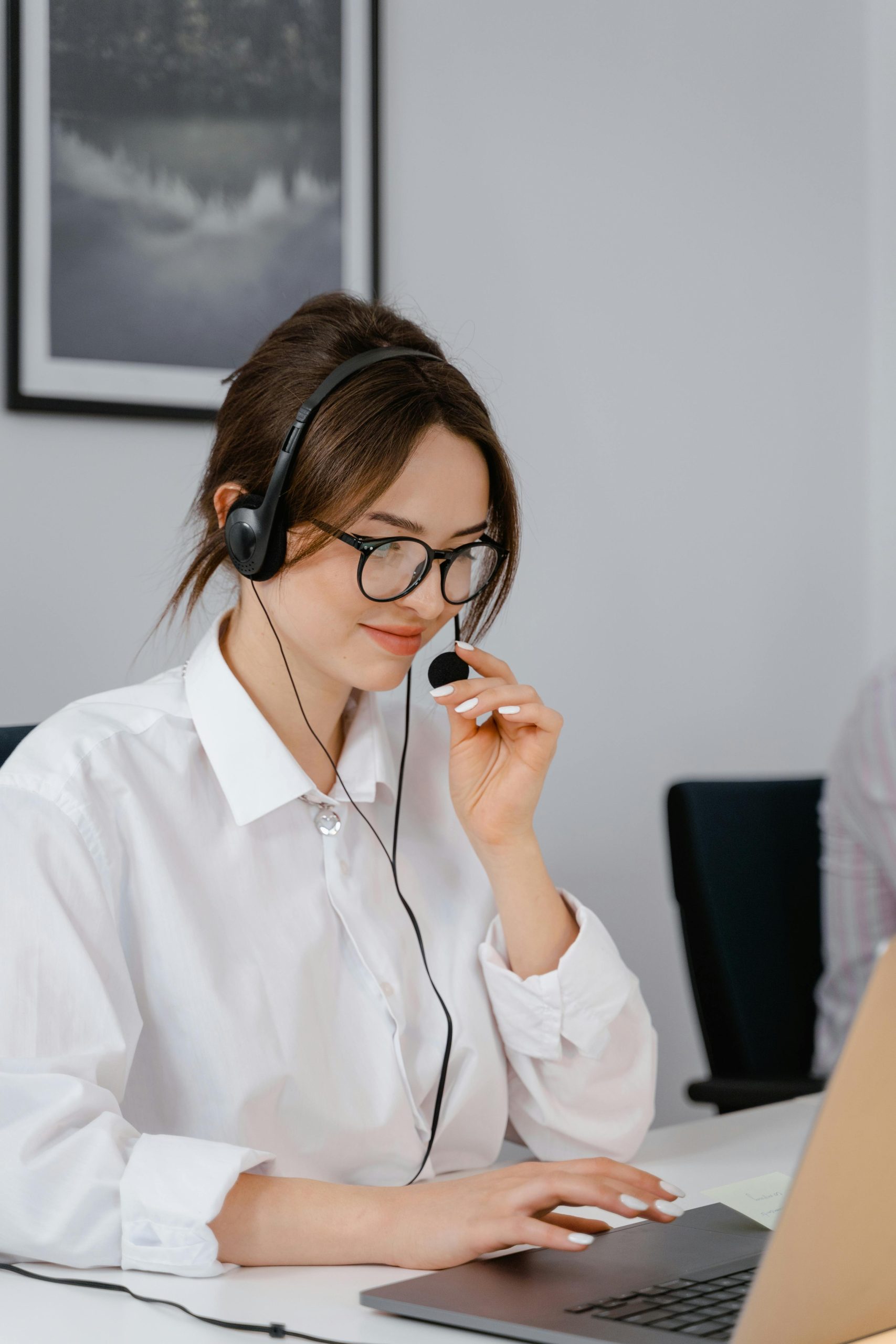 A woman in a white shirt and glasses is sitting at a desk, wearing a headset, looking at a laptop, and speaking into the microphone. A framed picture is on the wall behind her.