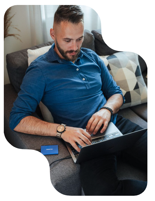 Man sitting on a couch, wearing a blue shirt and typing on a laptop. A portable hard drive rests beside him.