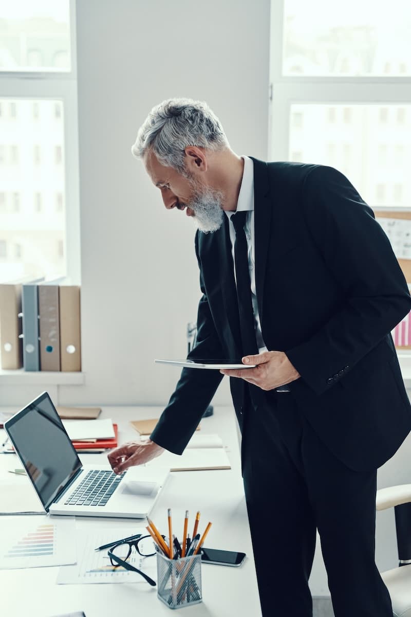 Elderly man in a suit using a laptop at a desk, holding a tablet. Office supplies are visible around him.