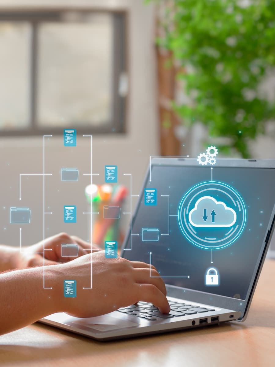 Person typing on a laptop displaying a cloud computing graphic with folders and data transfer symbols, set in a bright room with a plant.