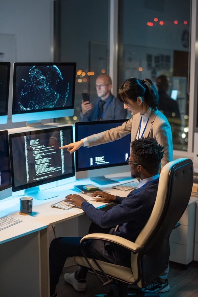 A woman points at a computer screen displaying code while a man sits at the desk. Another person uses a phone in the background. Multiple monitors and office equipment are visible.