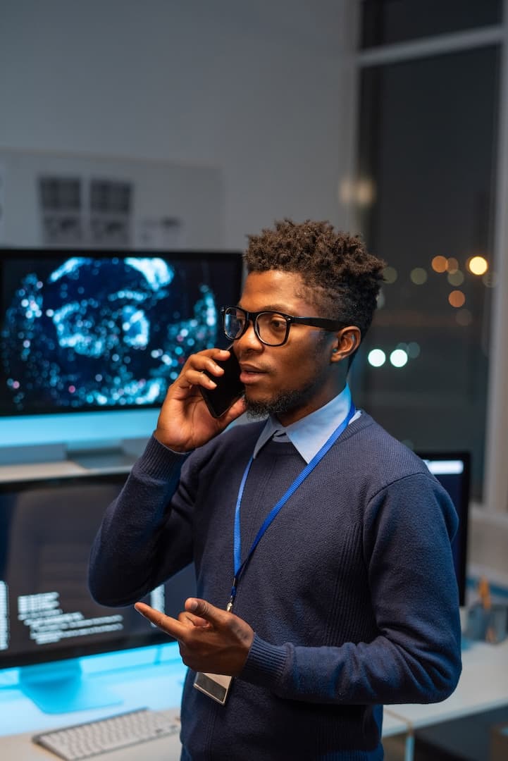 A man in a blue sweater talks on a smartphone in an office, standing in front of computer screens with digital displays.