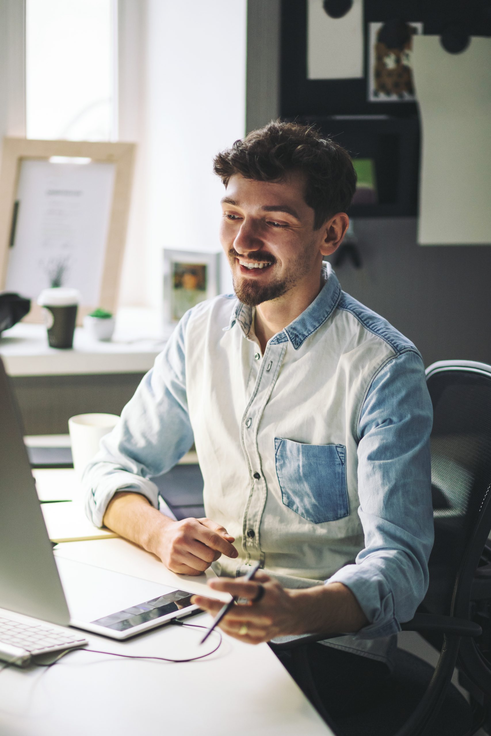 A man in a denim shirt smiles while sitting at his desk with a computer and smartphone, basking in the bright office light. It's another productive day in Grand Junction.