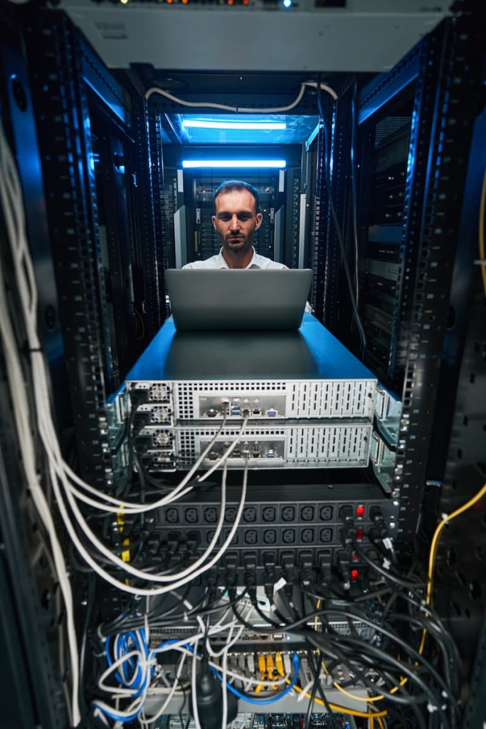 A technician works on a laptop in a server room surrounded by network equipment and cables.