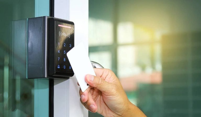 A hand holds a white card near a digital keypad access control system mounted on a glass door, all under the watchful eye of security camera systems.