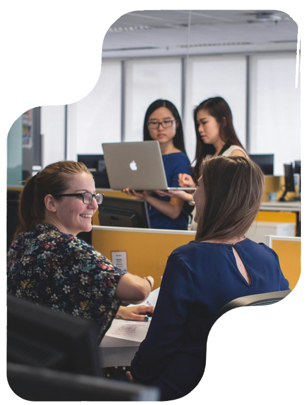 Four women in an office setting collaborate at desks, with two seated and two standing while holding a laptop.