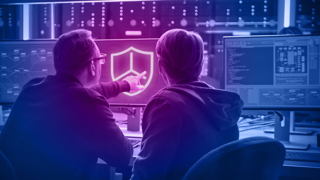 Two people sit at computer monitors in an office, with one person pointing at a screen displaying a large shield icon, suggesting cybersecurity or data protection work.