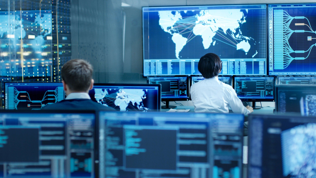 People work at computer stations in a control room, monitoring global network data displayed on large screens showing maps and digital information.