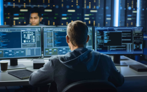 Person in hoodie sits at a desk working on multiple computer monitors displaying code and data, with a server room and another person visible in the background.